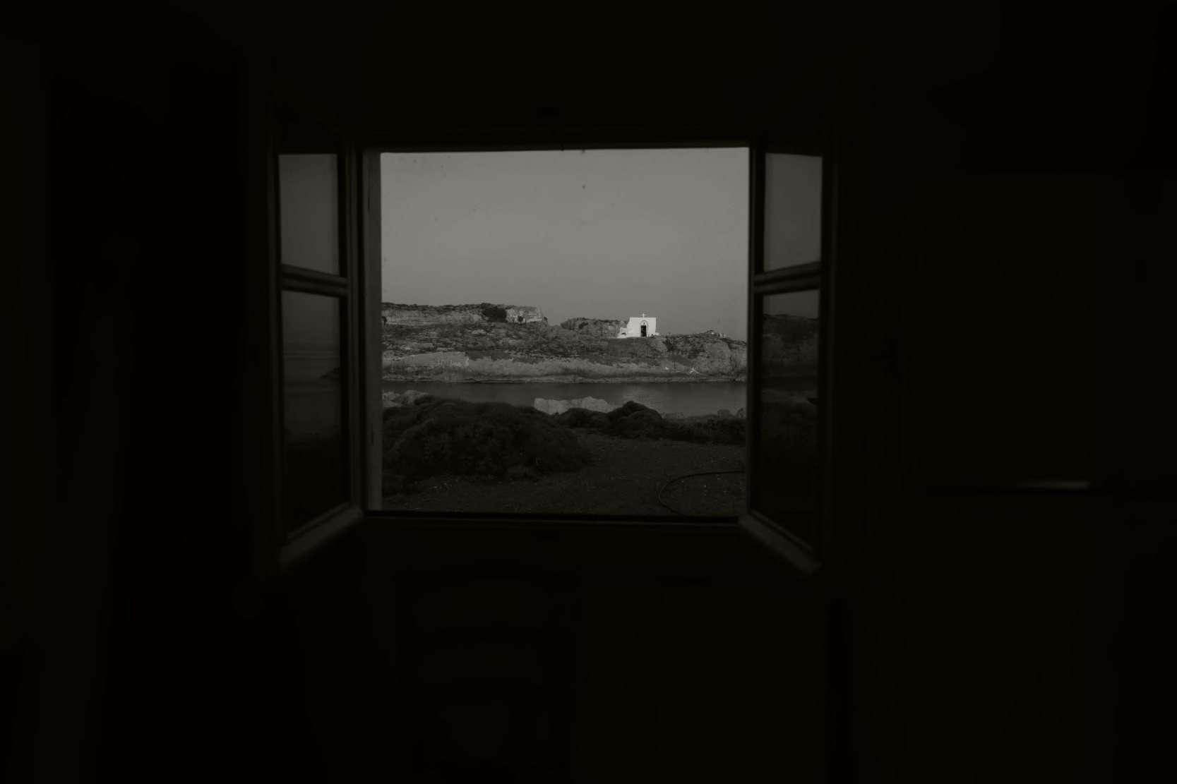 An open window in a dark room, looking out to a small white chapel by the sea