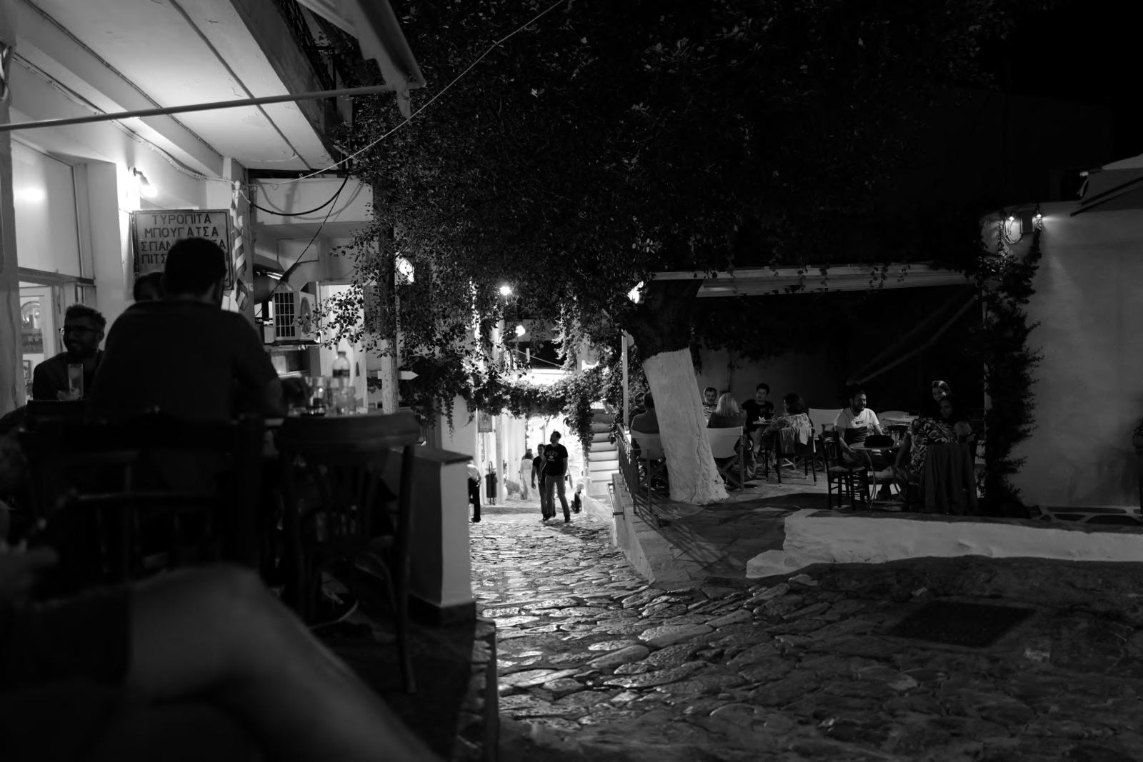 A cobbled street with café tables under a tree at night